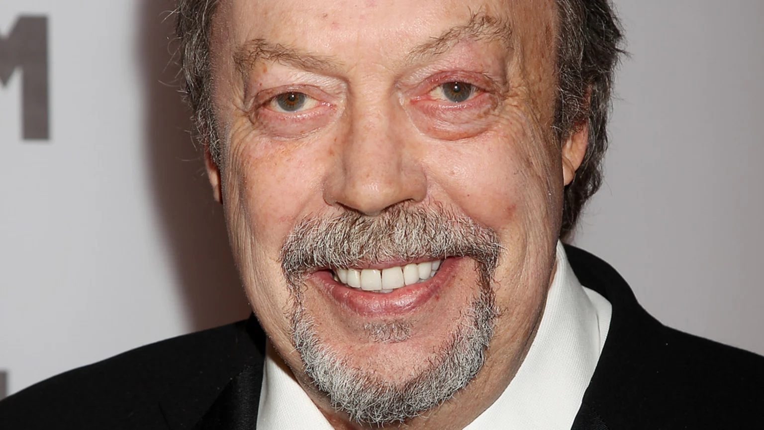Close-up of an older man with grey hair, mustache, and goatee, smiling while wearing a dark suit and white shirt.