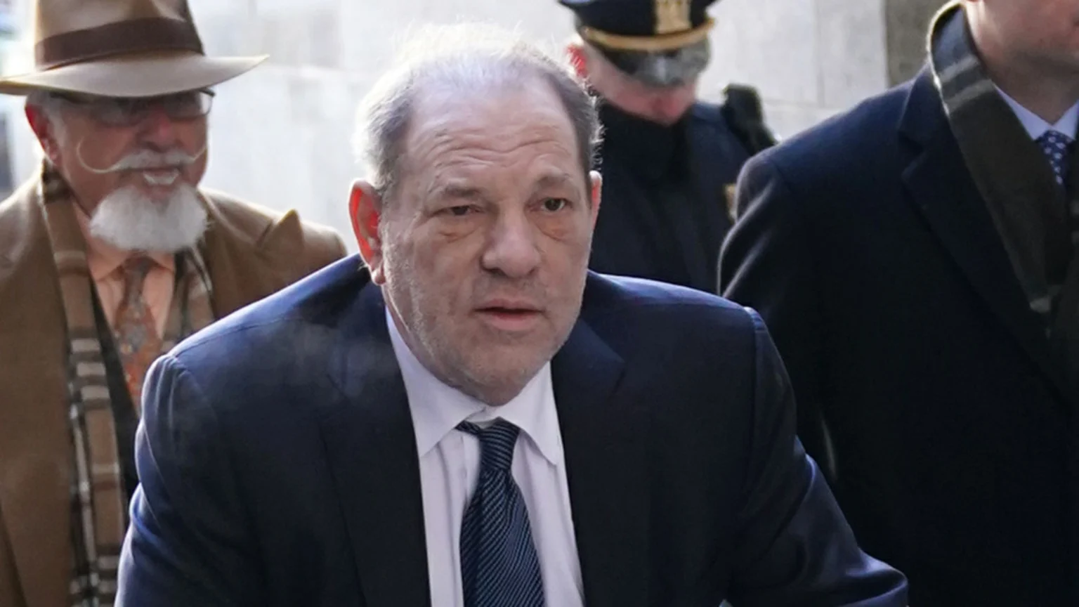 Harvey Weinstein in a dark suit and striped tie looks forward, with a man in a fedora and handlebar mustache smiling behind him.