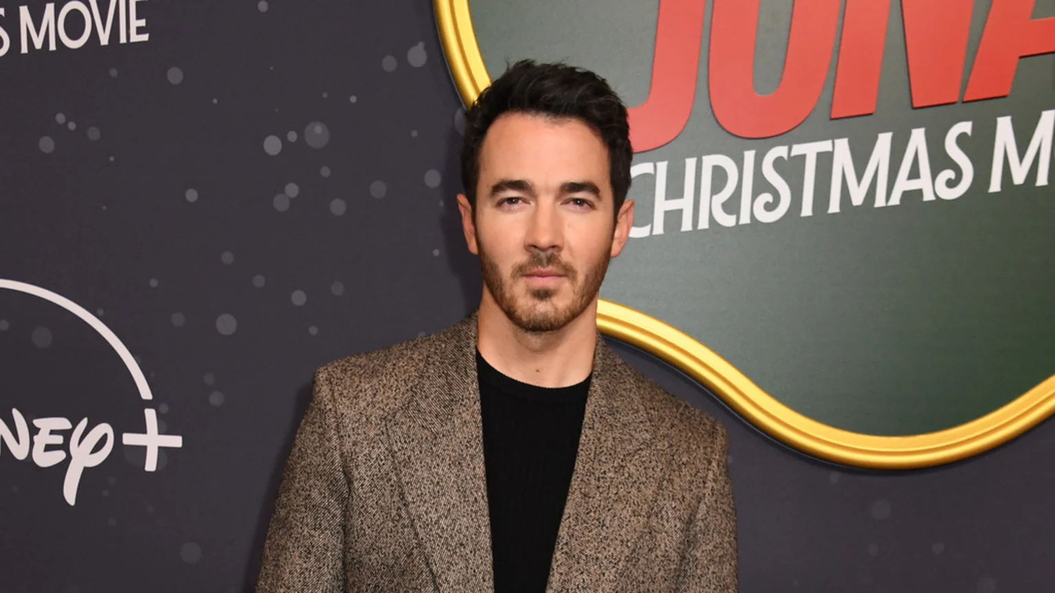 Kevin Jonas, a man with dark hair and a beard, looks at the camera wearing a black top and a brown blazer, standing against a dark background with Disney+ and "Jonas Christmas Movie" signage.