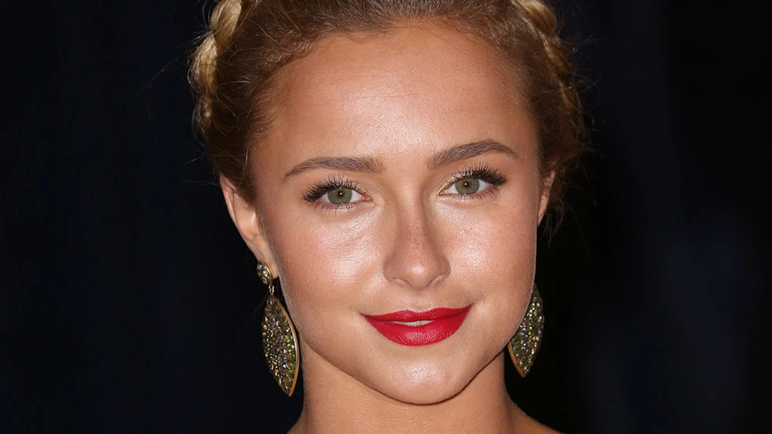 A woman with light brown braided hair, green eyes, red lipstick, and large ornate gold earrings with green gems looks directly at the camera.