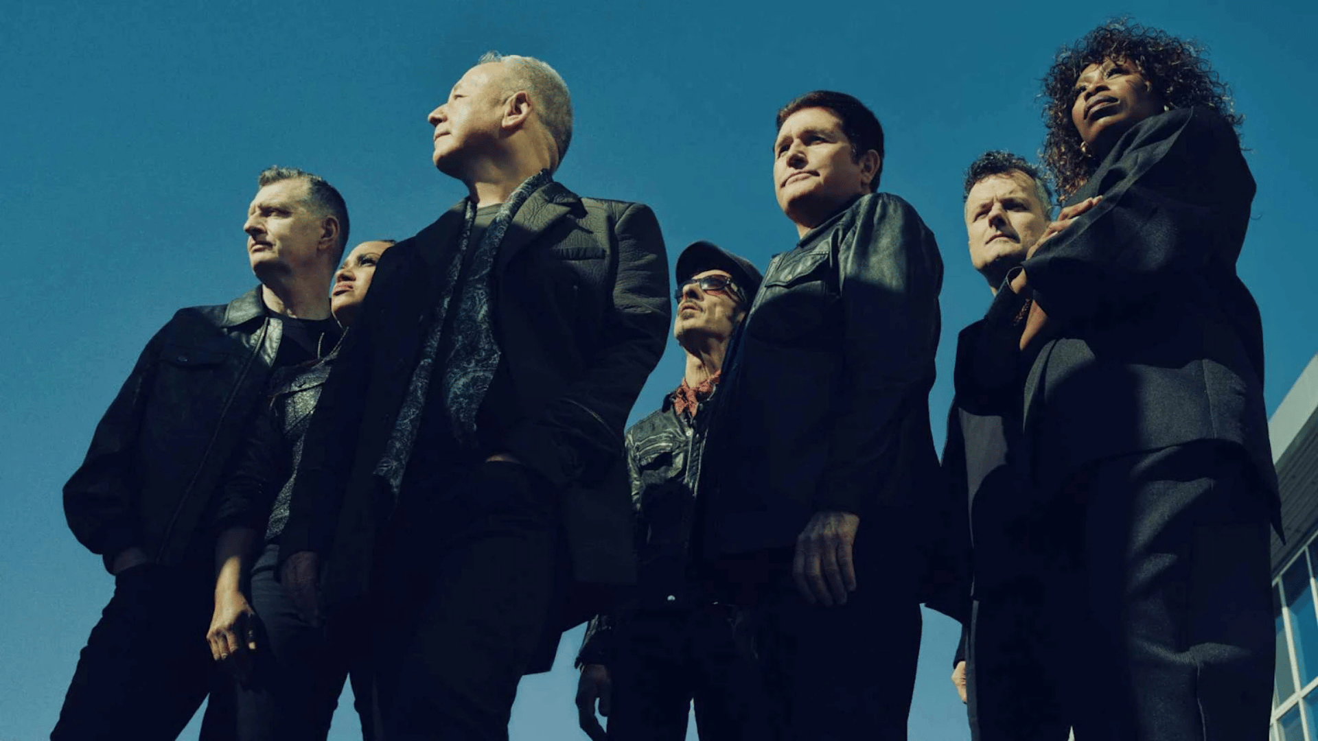 A group of seven adults, six men and one woman, stand outdoors looking up against a clear blue sky, dressed in dark jackets and suits, viewed from a low angle.
