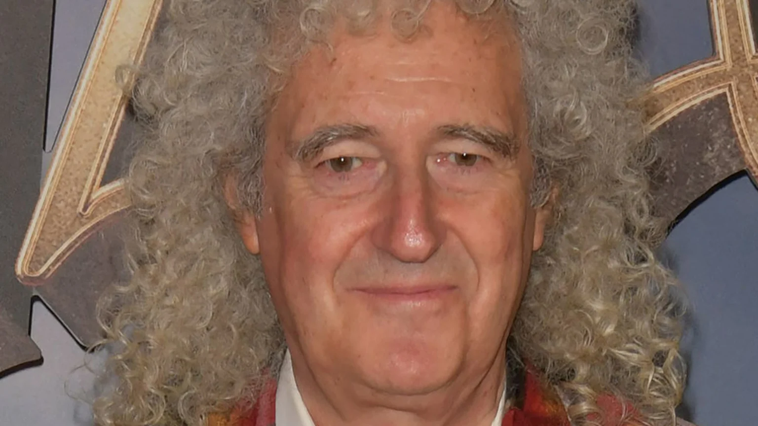 Close-up of an older man with curly grey-blonde hair, smiling slightly and looking forward, with parts of golden decorative elements in the background.