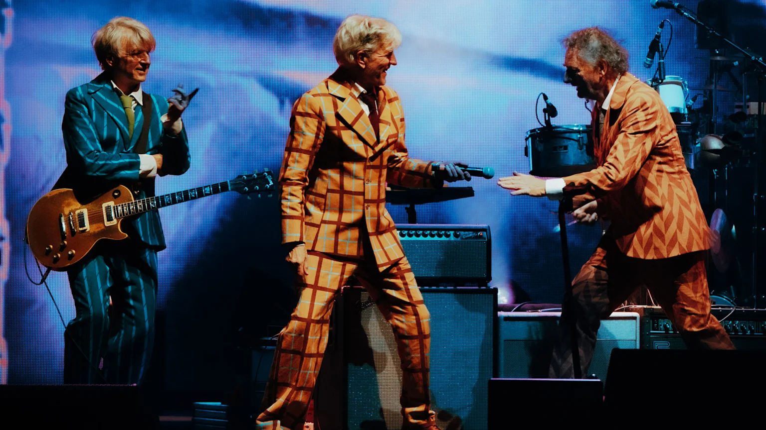 Three smiling musicians on stage, dressed in colorful patterned suits; one holds a guitar, and another extends a microphone to the third.