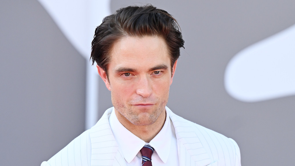 A close-up of actor Robert Pattinson shows him wearing a white pinstripe suit, a white shirt, and a striped tie, looking directly at the camera with dark, wavy hair and light stubble.