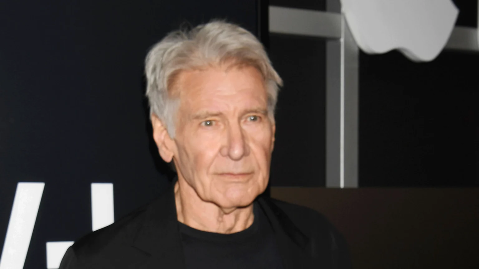 Harrison Ford with grey hair looks ahead, wearing a black top, against a dark background with a partial white Apple logo and other white shapes.