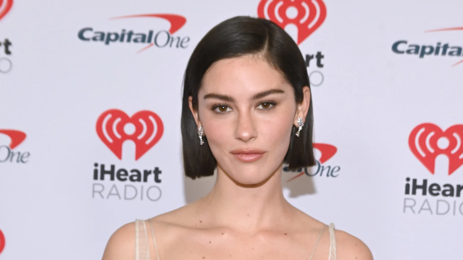 A woman with dark bobbed hair, brown eyes, and light makeup wears a light-colored top and earrings, standing against a white backdrop with Capital One and iHeart Radio logos.