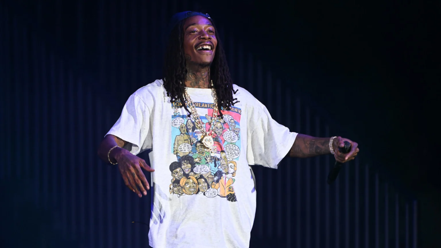 Wiz Khalifa smiles broadly on stage, wearing a blue baseball cap backward, dreadlocks, multiple gold chains, and an oversized light grey t-shirt with a colorful comic strip graphic, holding a microphone.
