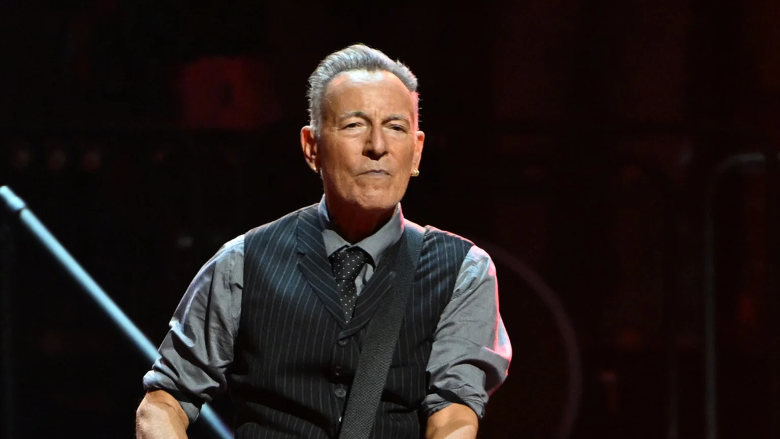 Bruce Springsteen, an older man with slicked-back grey hair, a serious expression, and gold earrings, wears a pinstriped vest, grey shirt, and patterned tie, with a guitar strap over his shoulder.
