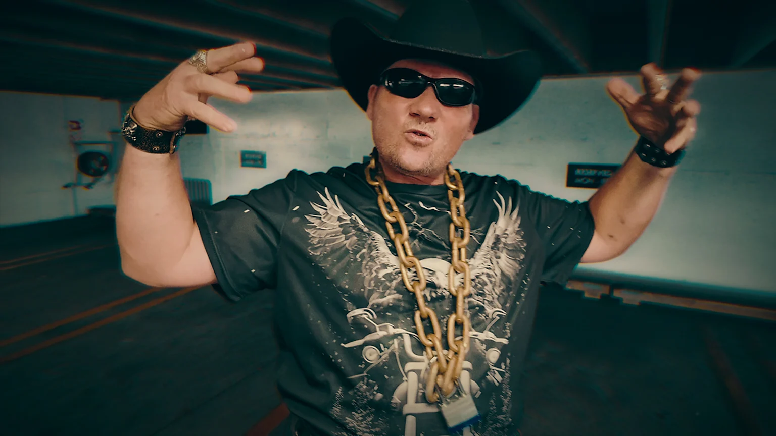 A man in a black cowboy hat and sunglasses wears a dark graphic shirt featuring a skull, eagles, and a motorcycle, adorned with a thick gold chain, bracelets, and rings, posing with raised arms.