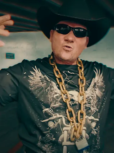 A man in a black cowboy hat and sunglasses wears a dark graphic shirt featuring a skull, eagles, and a motorcycle, adorned with a thick gold chain, bracelets, and rings, posing with raised arms.