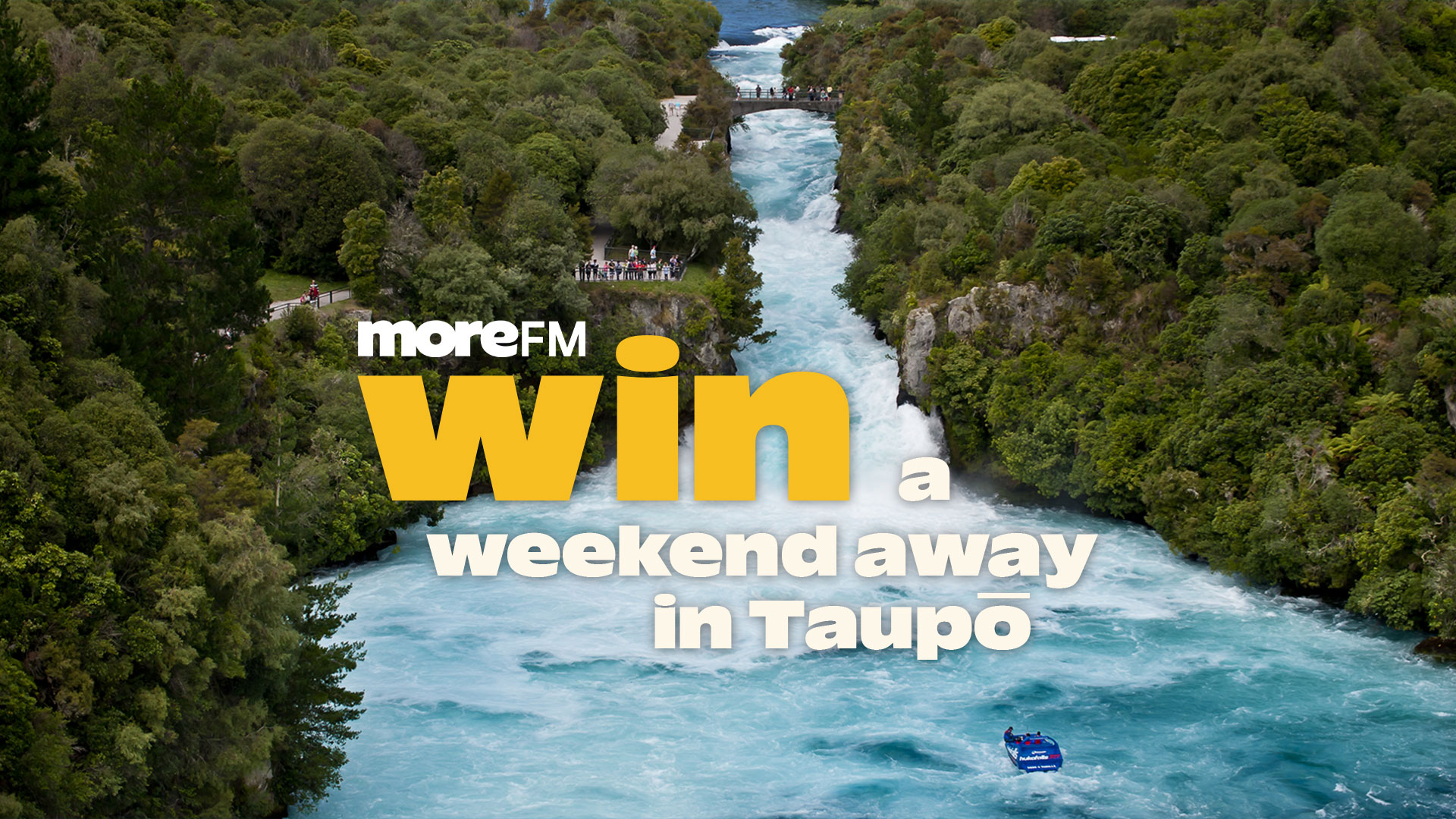 A river with rapids winds through a dense green forest, featuring a bridge, people, and a jet boat, with text: "moreFM Win a weekend away in Taupō".