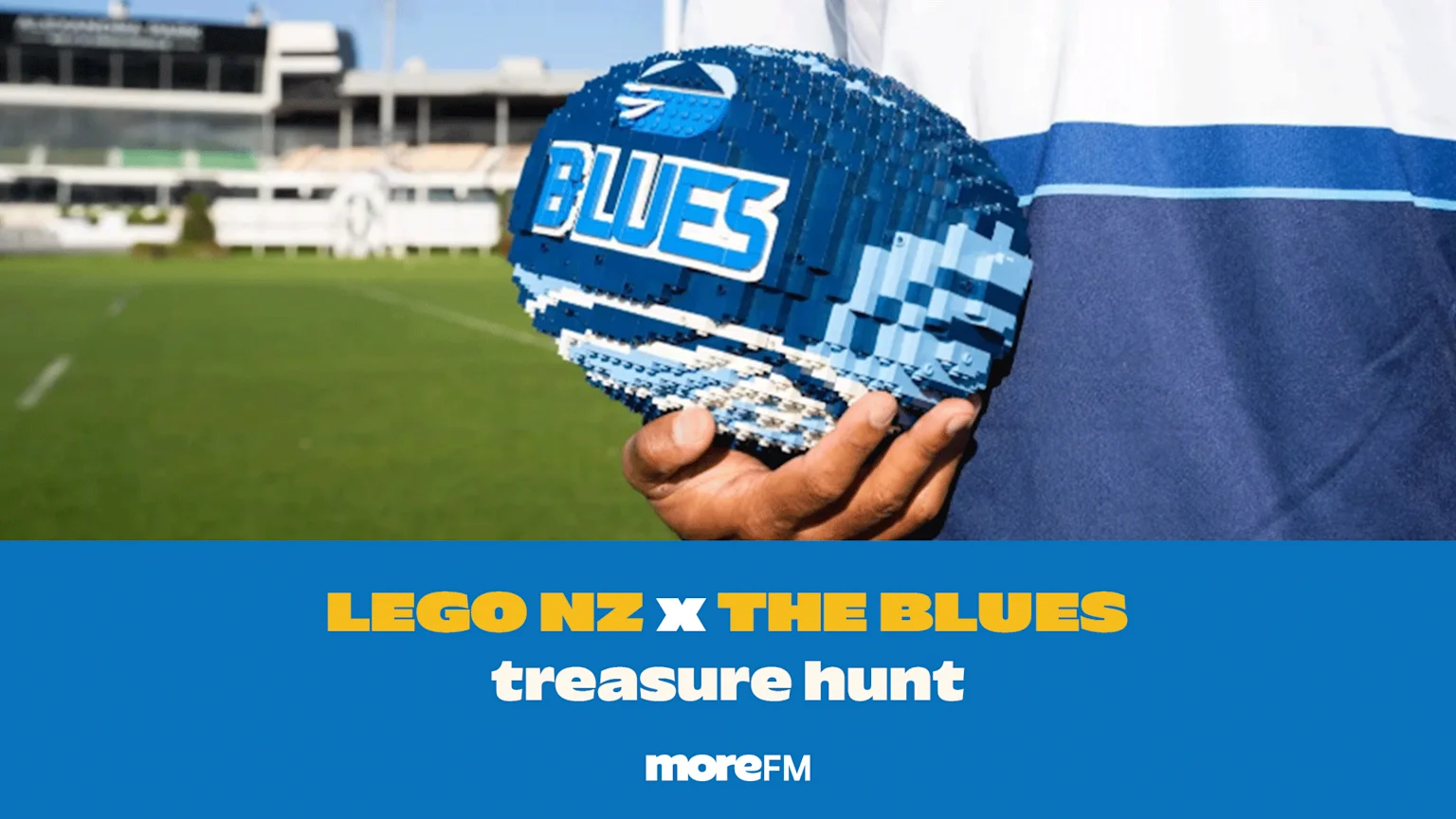 A person holds a blue and white LEGO brick rugby ball featuring the "BLUES" logo, on a green field with stadium seating; a blue banner beneath reads "LEGO NZ X THE BLUES treasure hunt moreFM".