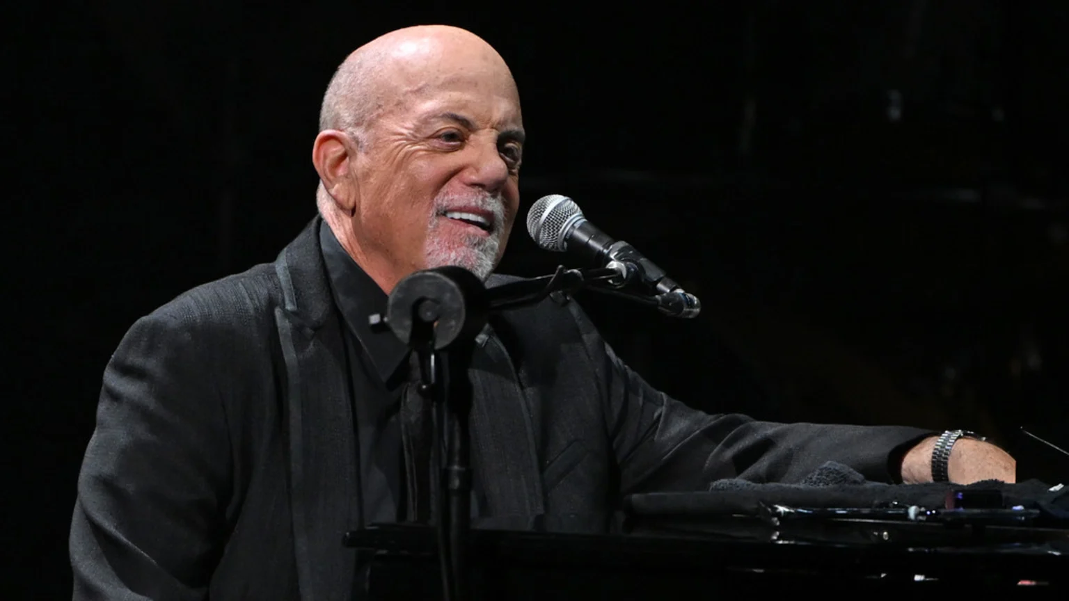 Billy Joel, with a bald head and grey beard, smiles widely while sitting at a piano with a microphone in front of him, wearing a dark suit.