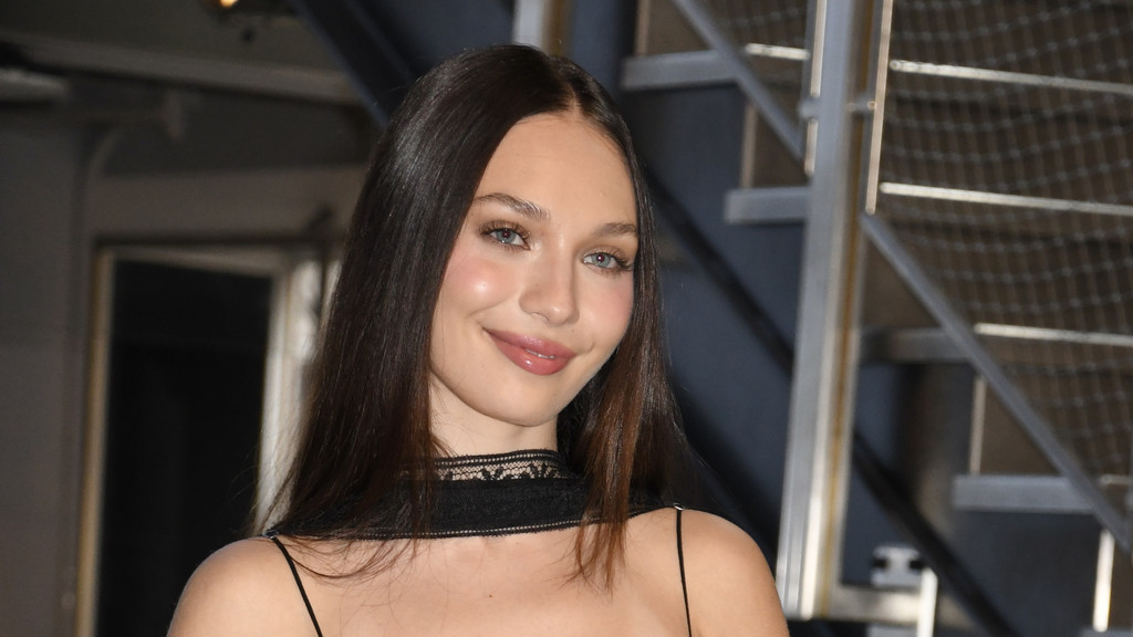 A young woman with long brown hair, blue eyes, and pink lips smiles softly, wearing a black spaghetti strap top with a lace choker, against a blurred background of metal stairs.