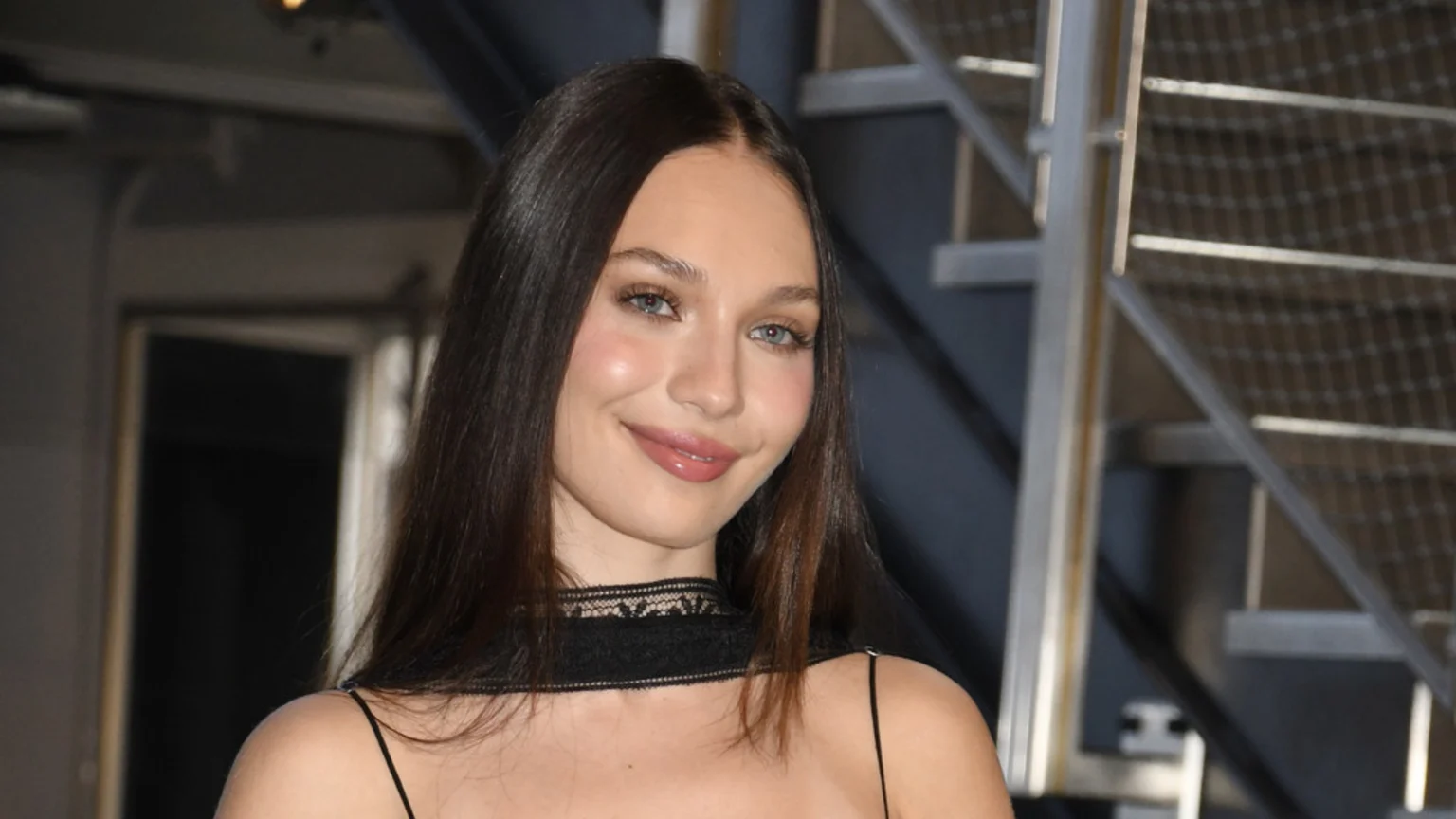 A young woman with long brown hair, blue eyes, and pink lips smiles softly, wearing a black spaghetti strap top with a lace choker, against a blurred background of metal stairs.