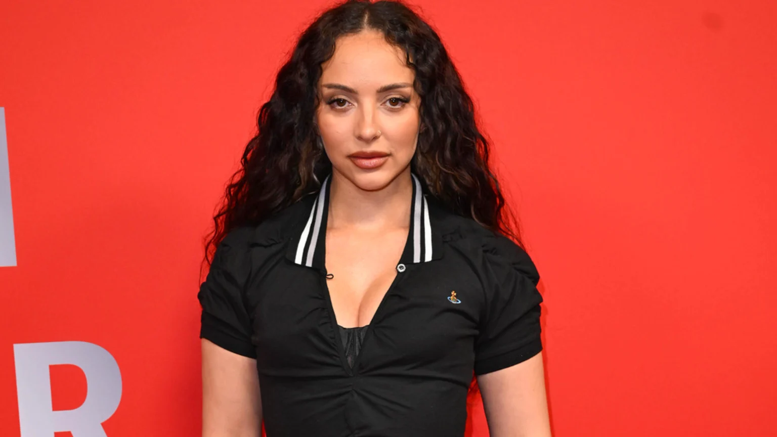 Jade Thirlwall looks at the camera, wearing a black collared shirt with a white striped collar and a black top underneath, against a red background with white text.