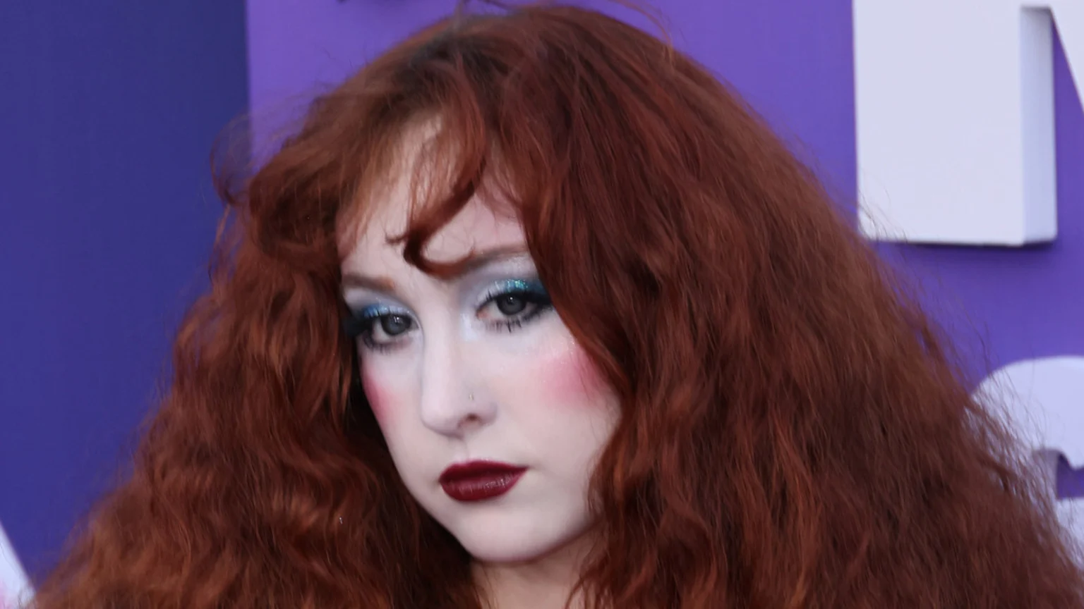 A close-up of a person with long, wavy red hair and dramatic makeup, including blue eyeshadow, pink blush, and dark red lipstick, against a blurry purple background.