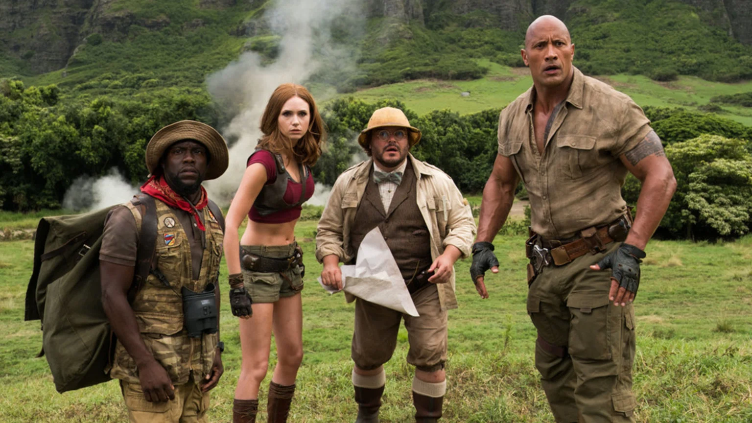Dwayne Johnson, Kevin Hart, Karen Gillan, and Jack Black stand in safari-style outfits in a jungle setting with mountains.