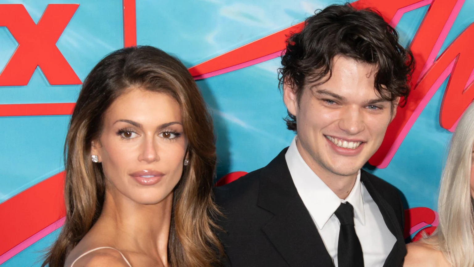 A smiling man with dark curly hair in a suit stands beside a woman with long brown hair wearing a light dress, both against a blue, red, and pink geometric background.