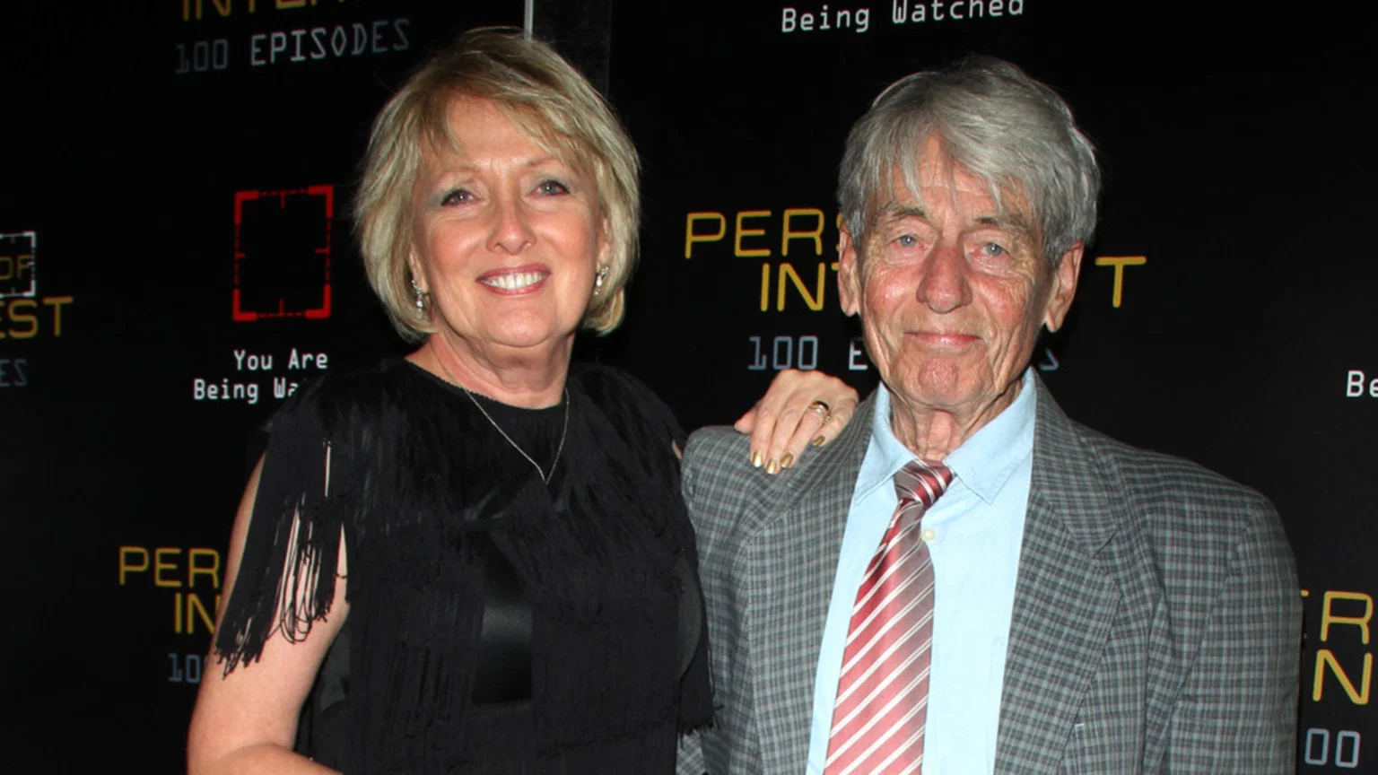 An older woman with blonde hair, gold nail polish, and a black fringe top smiles with her hand on the shoulder of an older man with grey hair, a plaid jacket, and a striped tie, in front of a dark "PERSON OF INTEREST" backdrop.