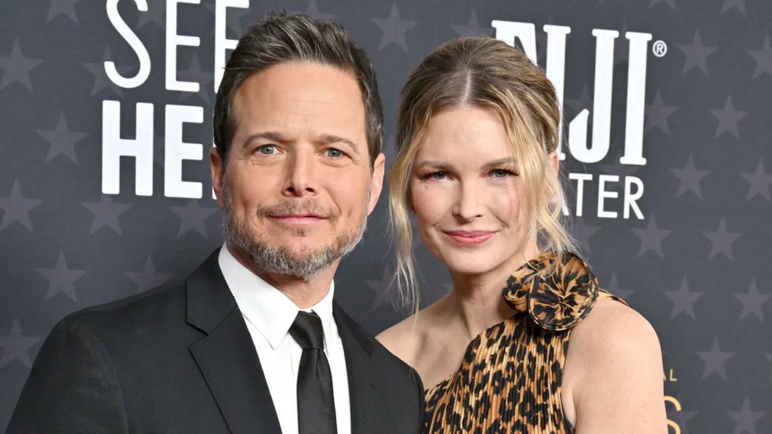 A man with a beard in a black suit and a blonde woman in a pleated leopard print dress pose together, smiling slightly against a dark background with stars and white text.