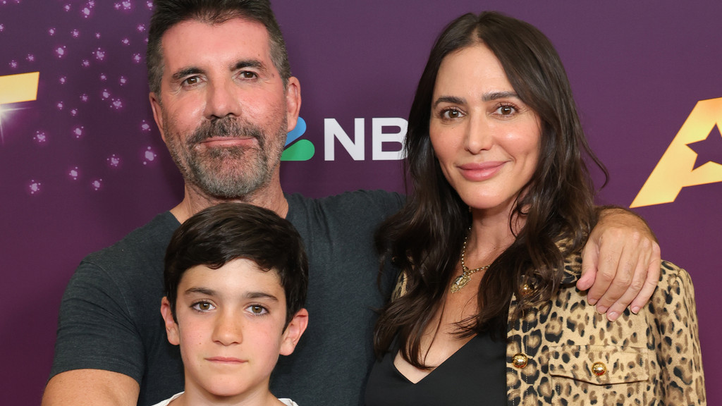 Simon Cowell, Lauren Silverman, and a young boy pose for a photo in front of a purple NBC banner.