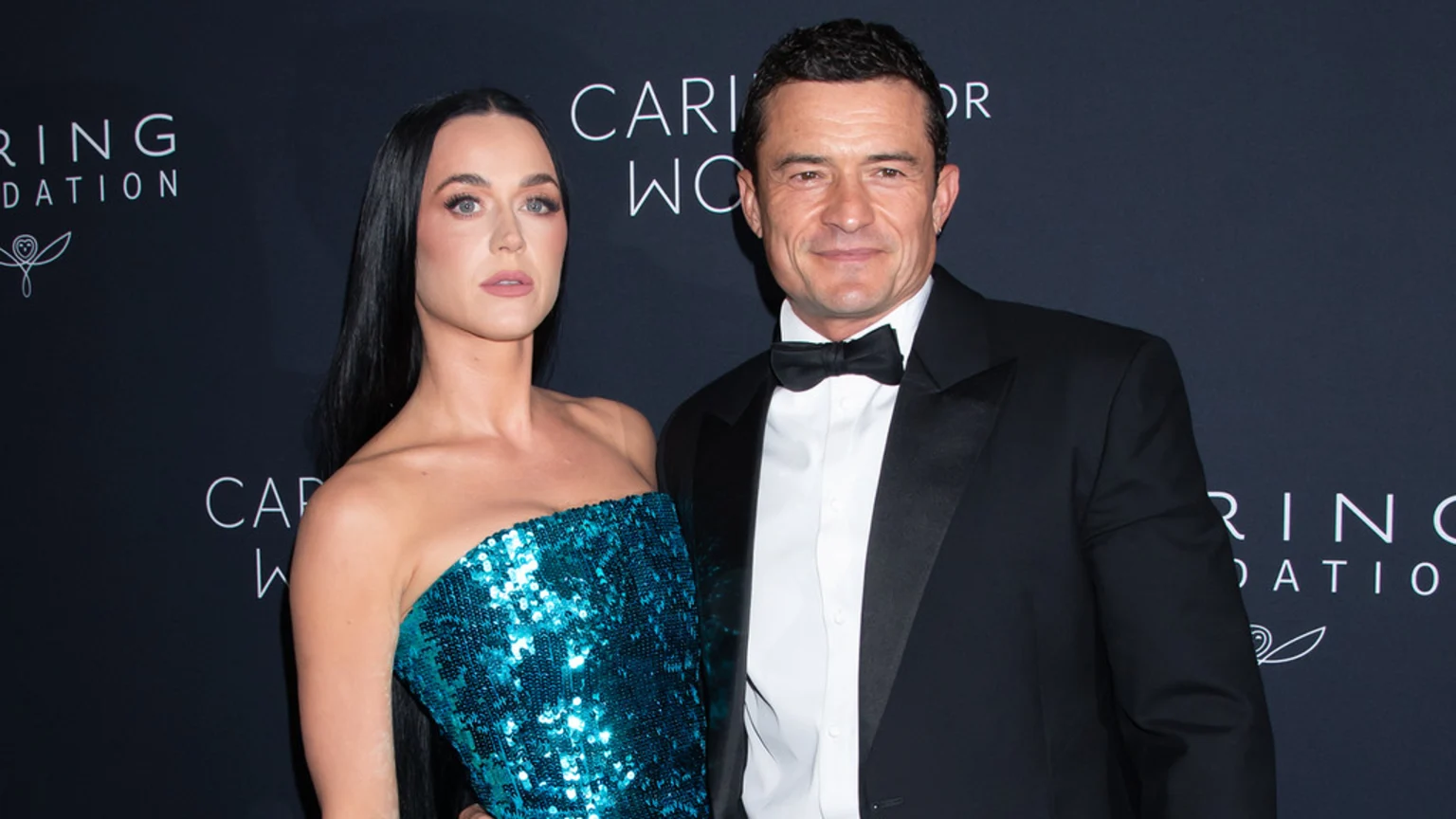 Katy Perry, in a strapless teal sequined dress, stands next to Orlando Bloom, who wears a black tuxedo, both looking at the camera against a dark event backdrop.