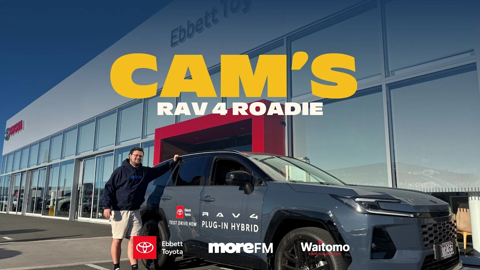A man stands in front of an Ebbett Toyota dealership next to a dark grey Toyota RAV4 Plug-in Hybrid branded with "Ebbett Toyota", "Waitomo", and "moreFM", under the large yellow and white text "CAM'S RAV 4 ROADIE".