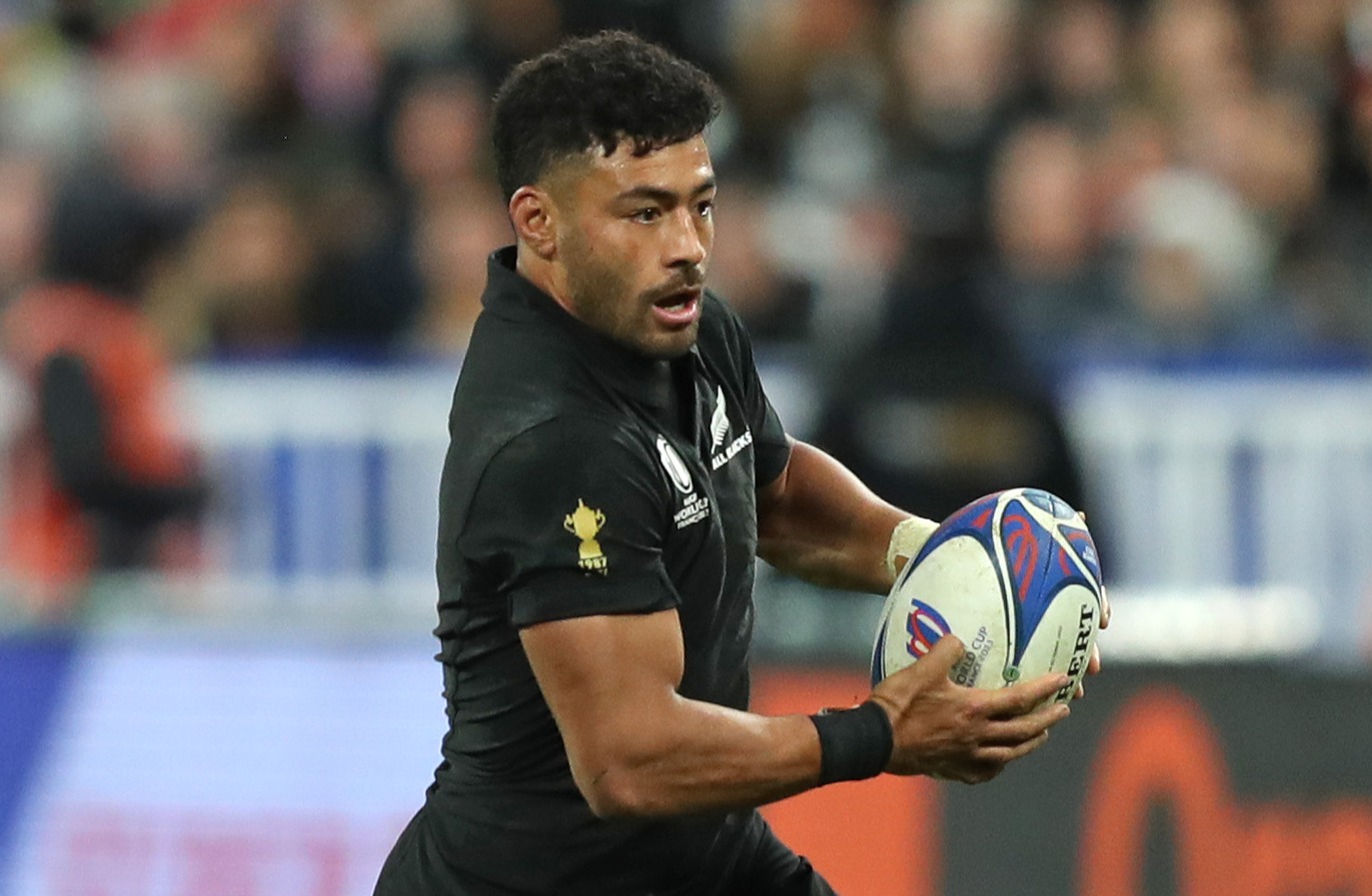A male rugby player in a black jersey with an All Blacks logo and a 1987 World Cup trophy emblem holds a rugby ball while looking forward during a game.