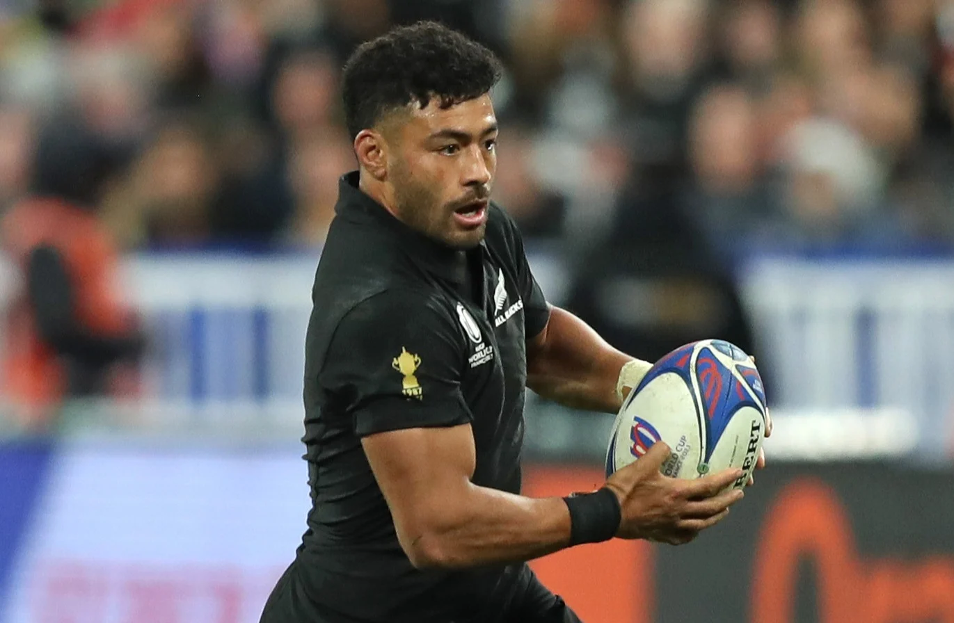 A male rugby player in a black jersey with an All Blacks logo and a 1987 World Cup trophy emblem holds a rugby ball while looking forward during a game.