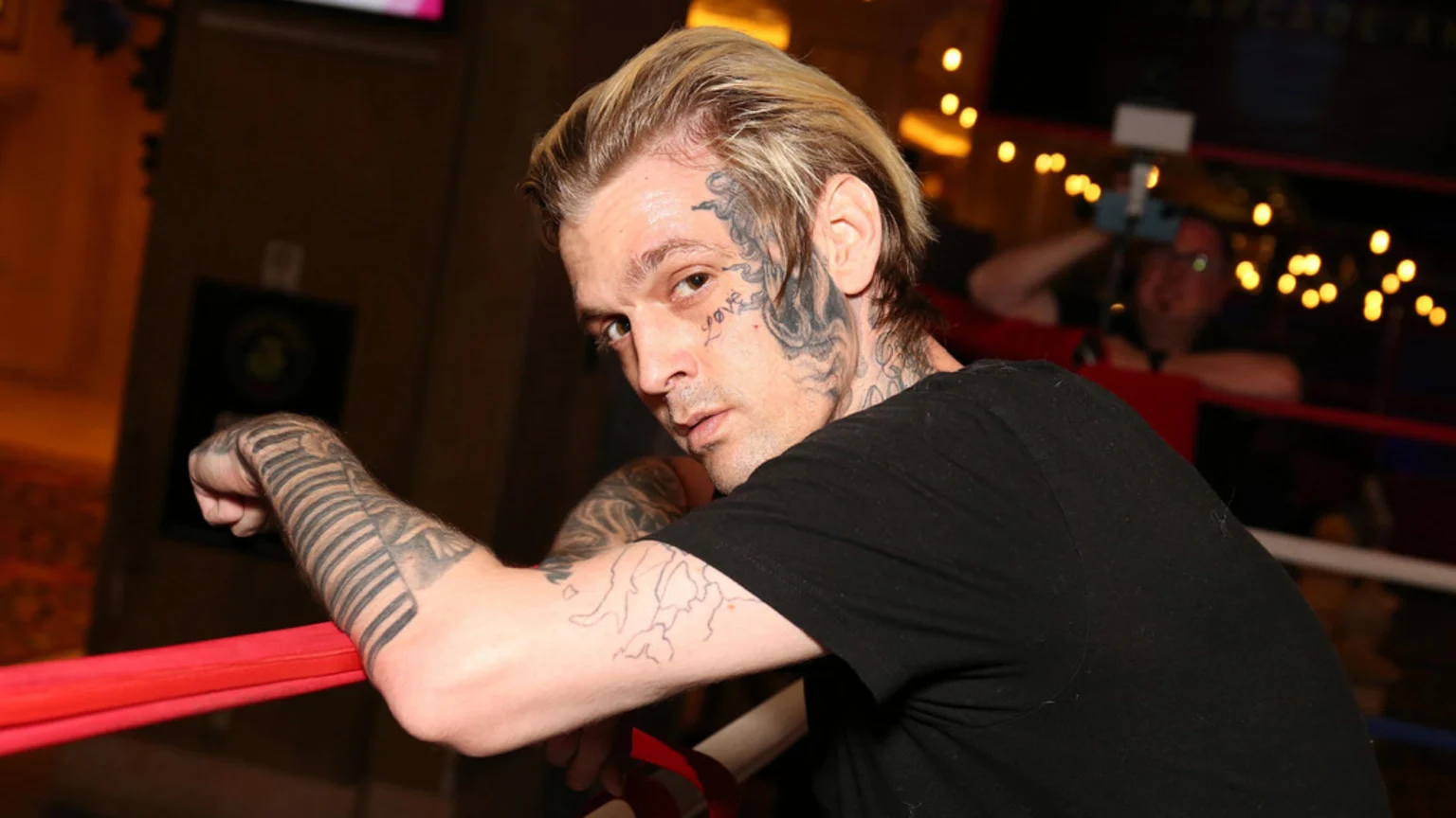 Aaron Carter with blonde hair and face tattoos including "Love" near his eye, wearing a black t-shirt, leaning on a red rope and looking at the camera.