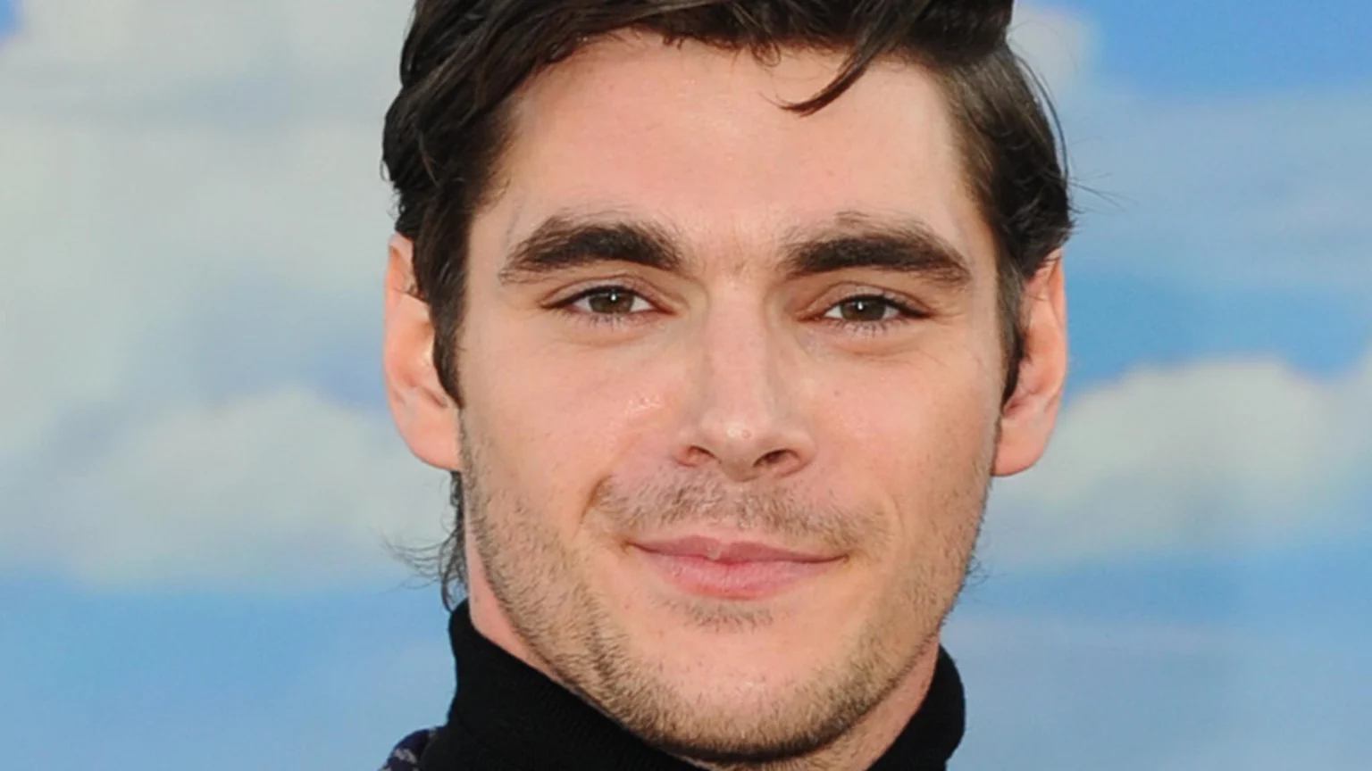 Close-up portrait of a young man with dark, wavy hair, prominent eyebrows, brown eyes, and light stubble, smiling subtly while wearing a black turtleneck against a blurry blue sky.