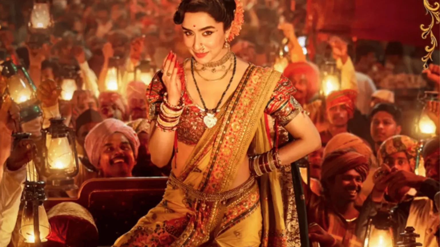 A smiling woman in a yellow saree, red patterned blouse, and traditional Indian jewelry sits, surrounded by a cheerful crowd holding glowing lanterns.