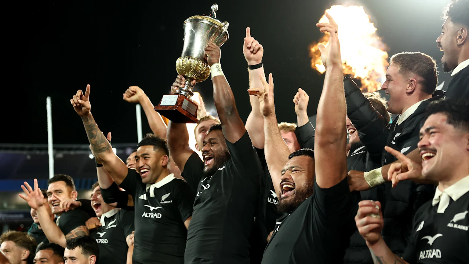 A group of jubilant men in black jerseys with silver fern logos celebrate at night, one holding a silver trophy aloft, with a bright flame illuminating faces on the right.