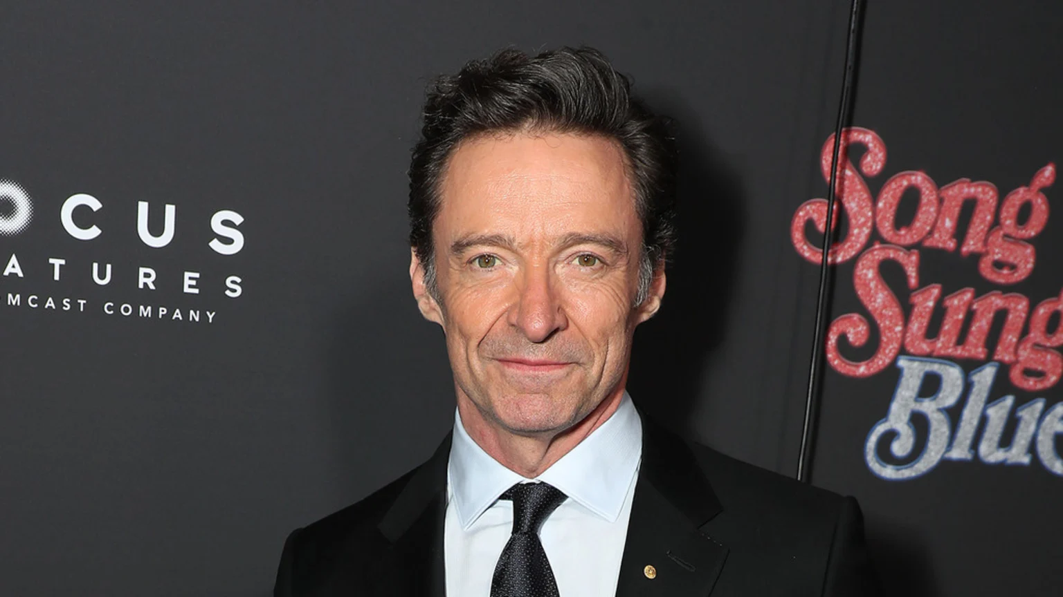 Hugh Jackman, in a black suit, white shirt, and black tie, smiles in front of a black backdrop featuring "FOCUS FEATURES" and glittery "Song Sung Blue" text.