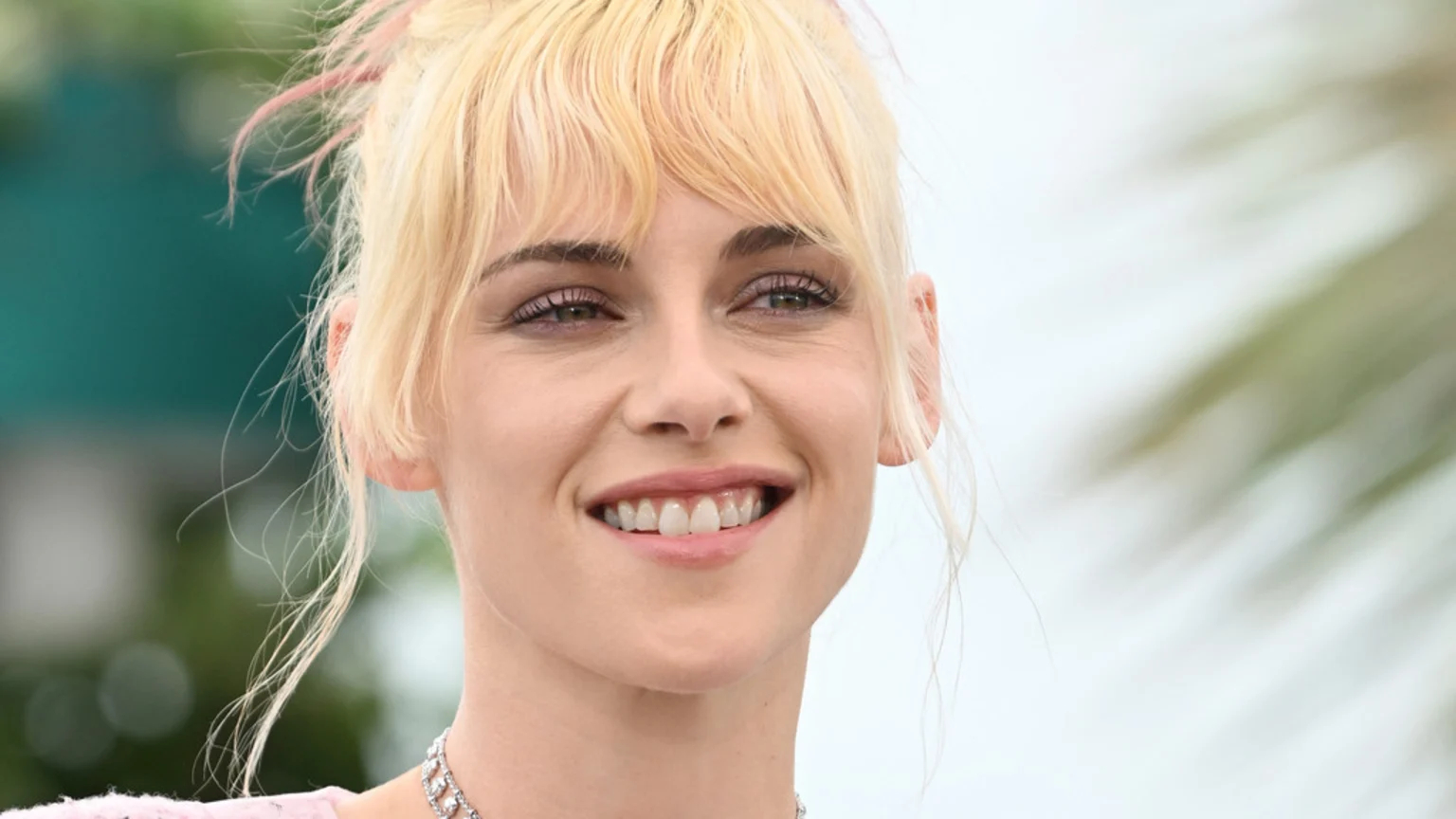 Close-up of Kristen Stewart smiling, wearing a silver necklace, with blonde hair and bangs, and a blurred green and white background.