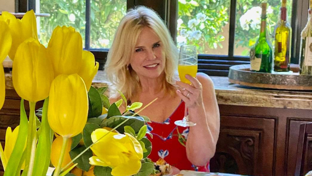 A smiling blonde woman in a red floral top holds a glass with a yellow drink, with yellow tulips in the foreground and wine bottles on a counter in the background.