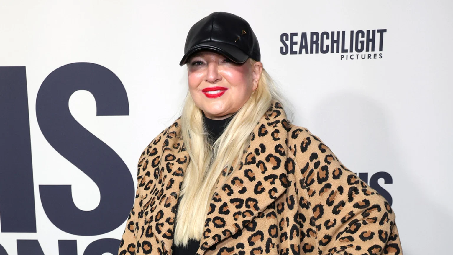 Singer Sia, with long blonde hair, red lipstick, a black leather cap, and a leopard print coat, smiles at the camera in front of a white background with "SEARCHLIGHT PICTURES" visible.