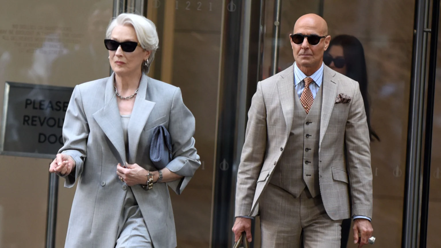 A woman with white hair and sunglasses in a light grey pantsuit walks past a glass door next to a bald man in a light brown plaid three-piece suit and sunglasses.