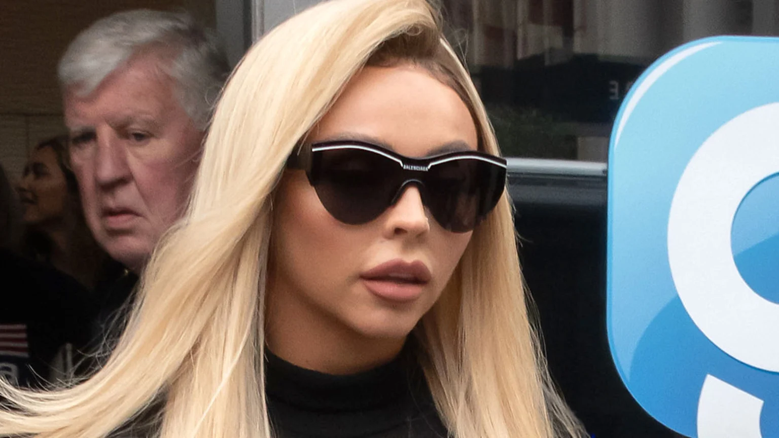 A woman with long blonde hair wears large Balenciaga sunglasses and a black top, looking forward, with a grey-haired man partially visible behind her.