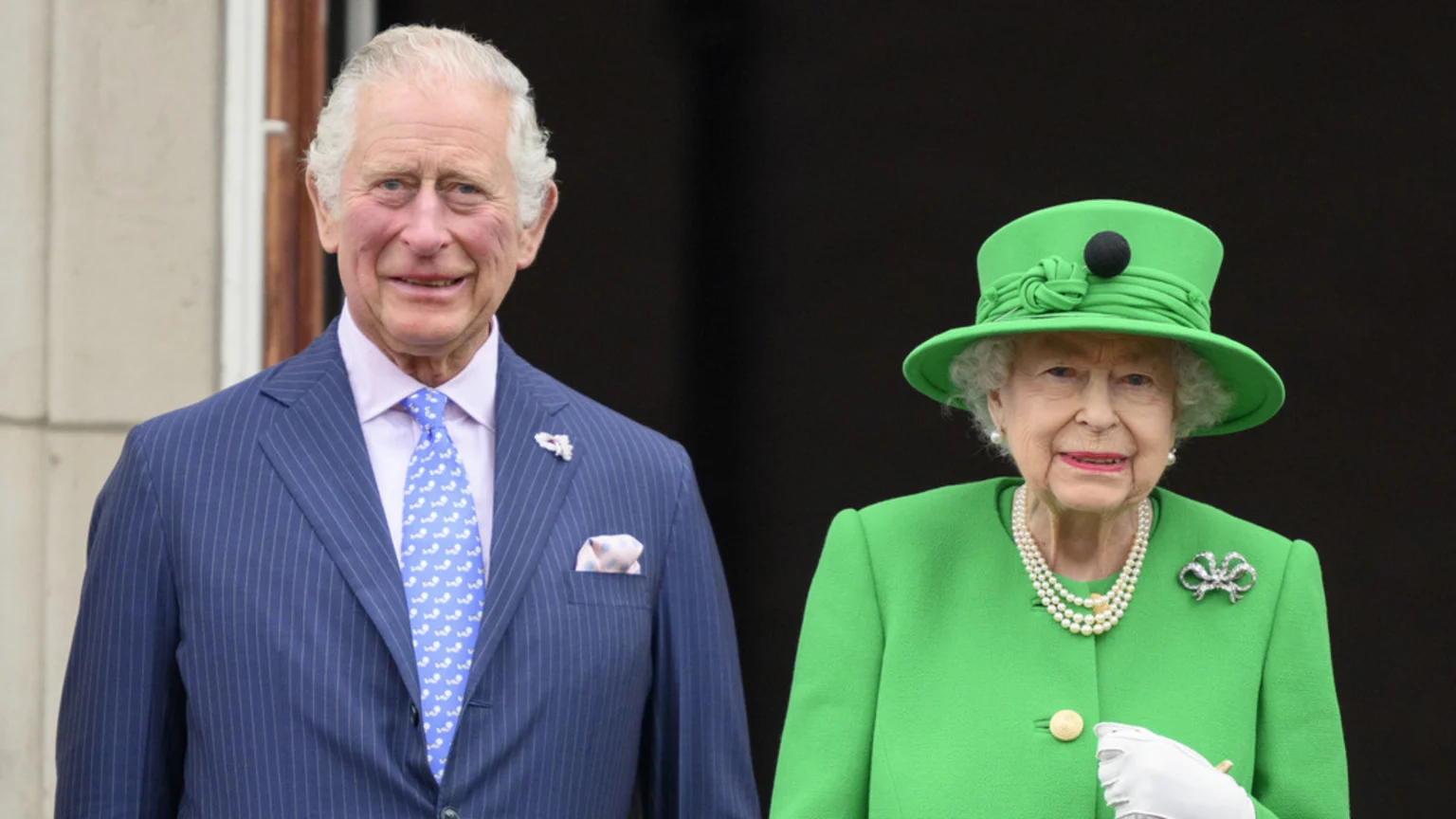 King Charles III in a blue pinstripe suit and Queen Elizabeth II in a bright green coat and hat stand together, smiling.