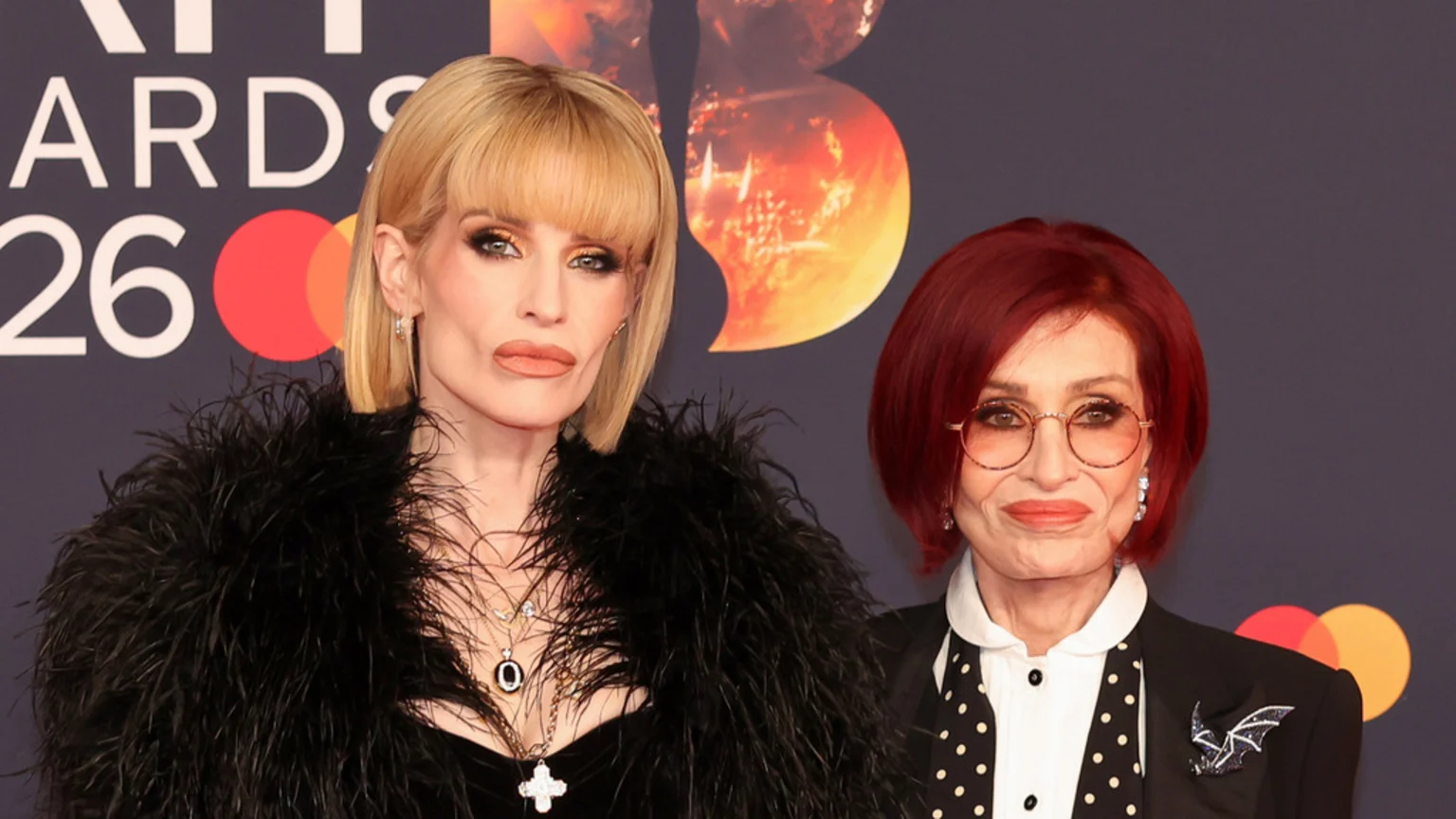 Kelly and Sharon Osbourne pose on a red carpet; Kelly, left, has blonde hair and a black feathered top, while Sharon, right, has red hair, round glasses, and a black blazer with a polka-dot tie.