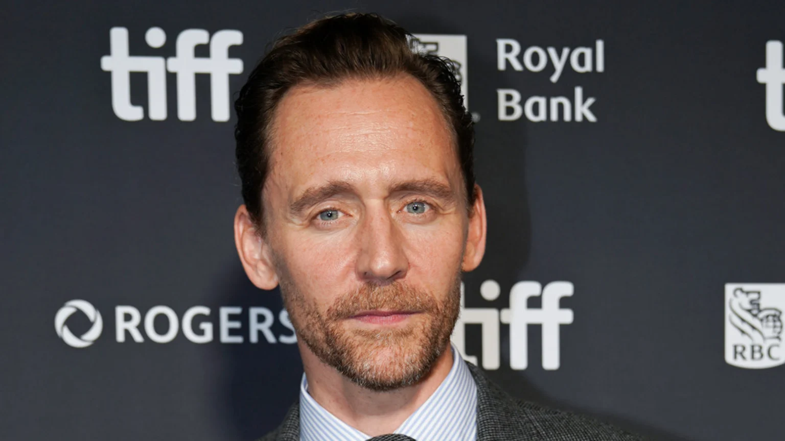 Actor Tom Hiddleston, with light blue eyes, brown hair, and a reddish beard, looks directly at the camera with a neutral expression, wearing a striped shirt and blazer against a dark background with sponsor logos.