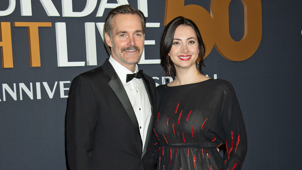 Actor Will Forte, in a tuxedo, stands smiling next to a woman in a dark dress with red accents, on a red carpet with a 'Saturday Night Live 50' backdrop.