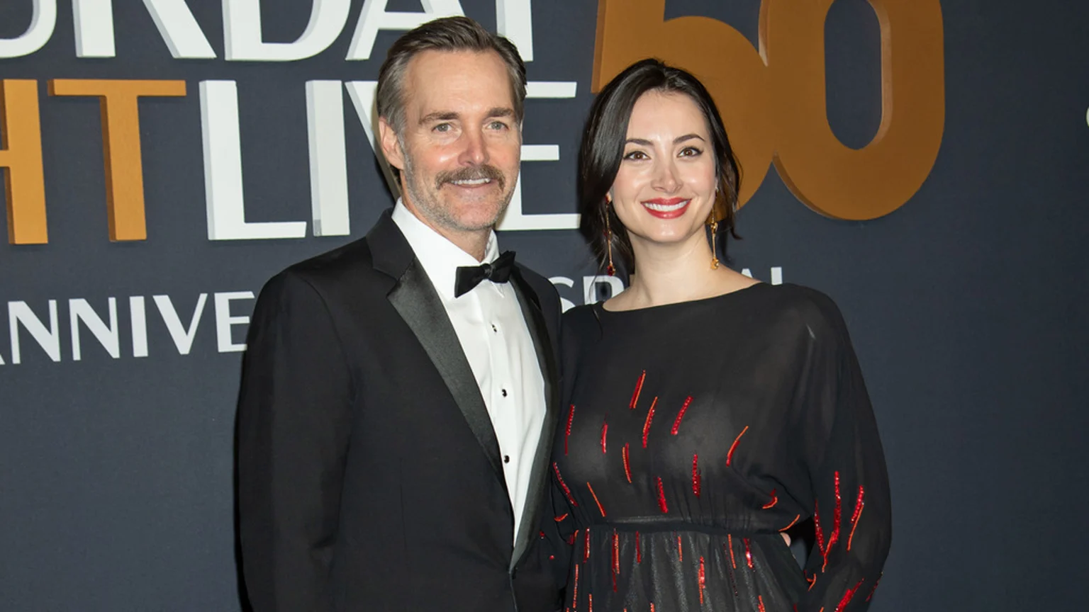 Actor Will Forte, in a tuxedo, stands smiling next to a woman in a dark dress with red accents, on a red carpet with a 'Saturday Night Live 50' backdrop.