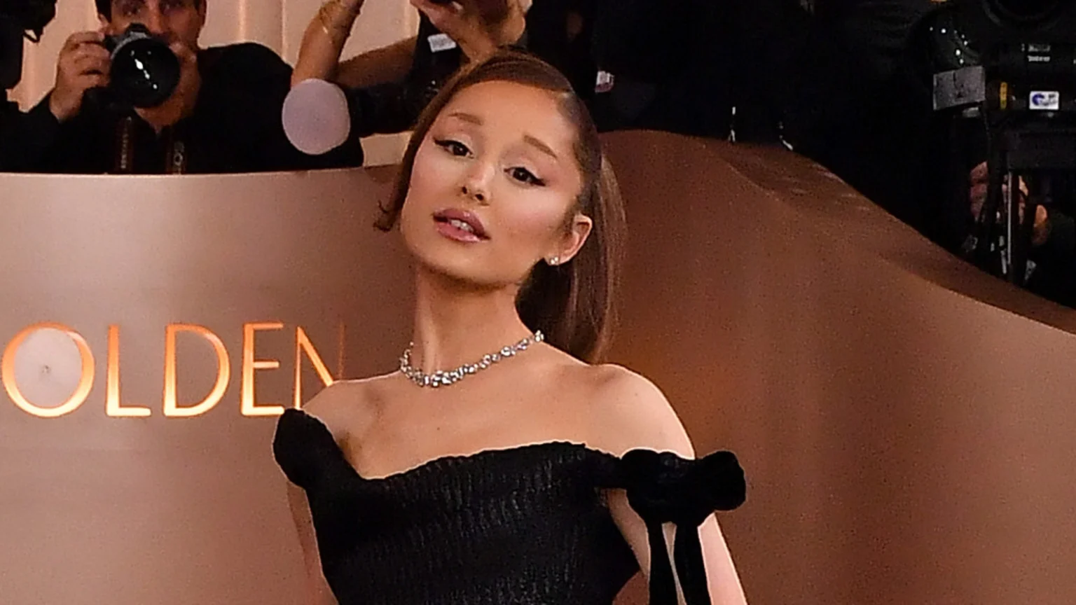 Ariana Grande poses for the camera in a black off-the-shoulder dress, a diamond necklace, and stud earrings, with her hair in a ponytail, against a light brown backdrop featuring the word "GOLDEN" and blurry figures.