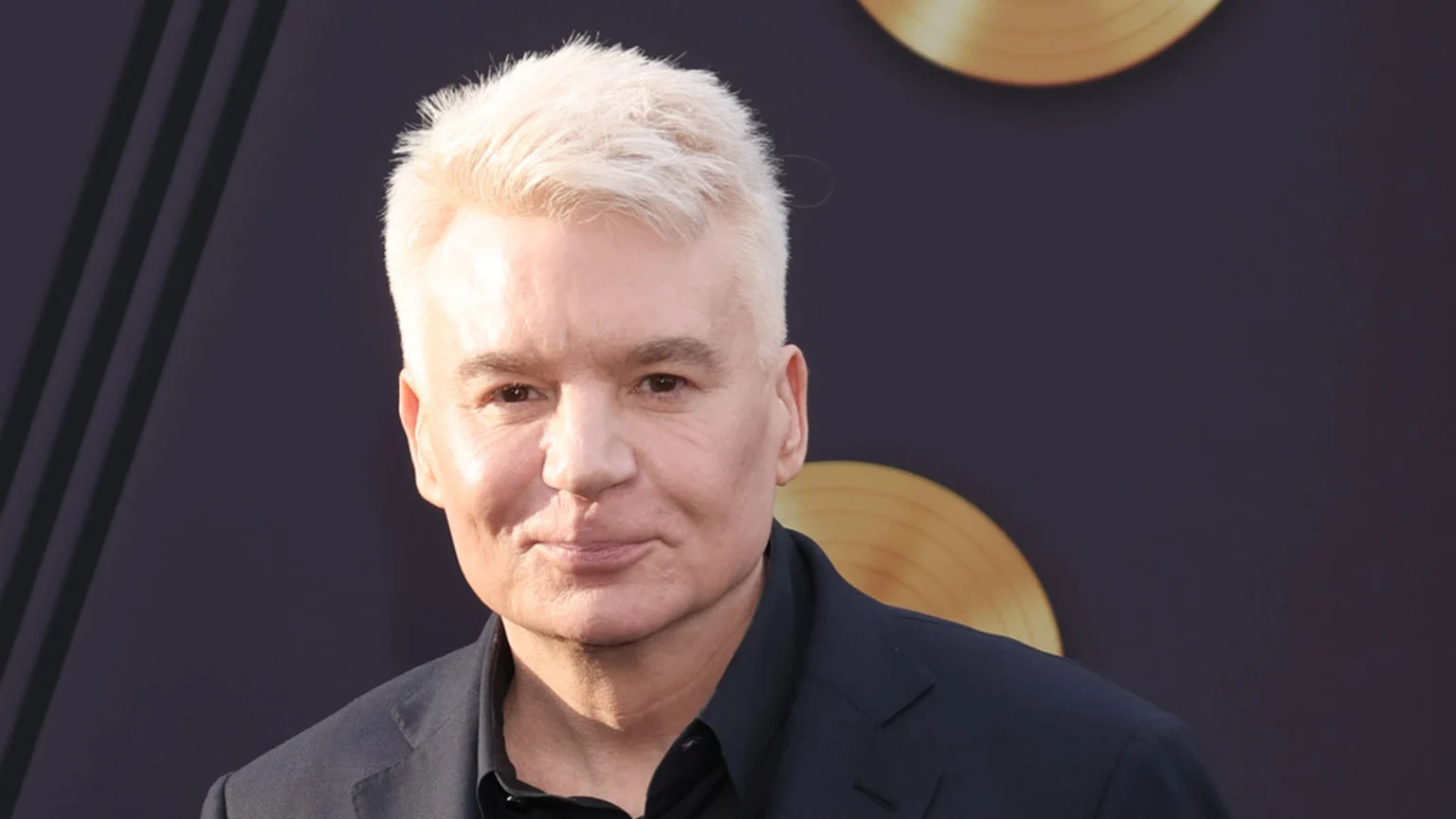 Close-up of actor Mike Myers with short, platinum blonde hair, wearing a dark suit and shirt, smiling subtly against a dark background with gold accents.