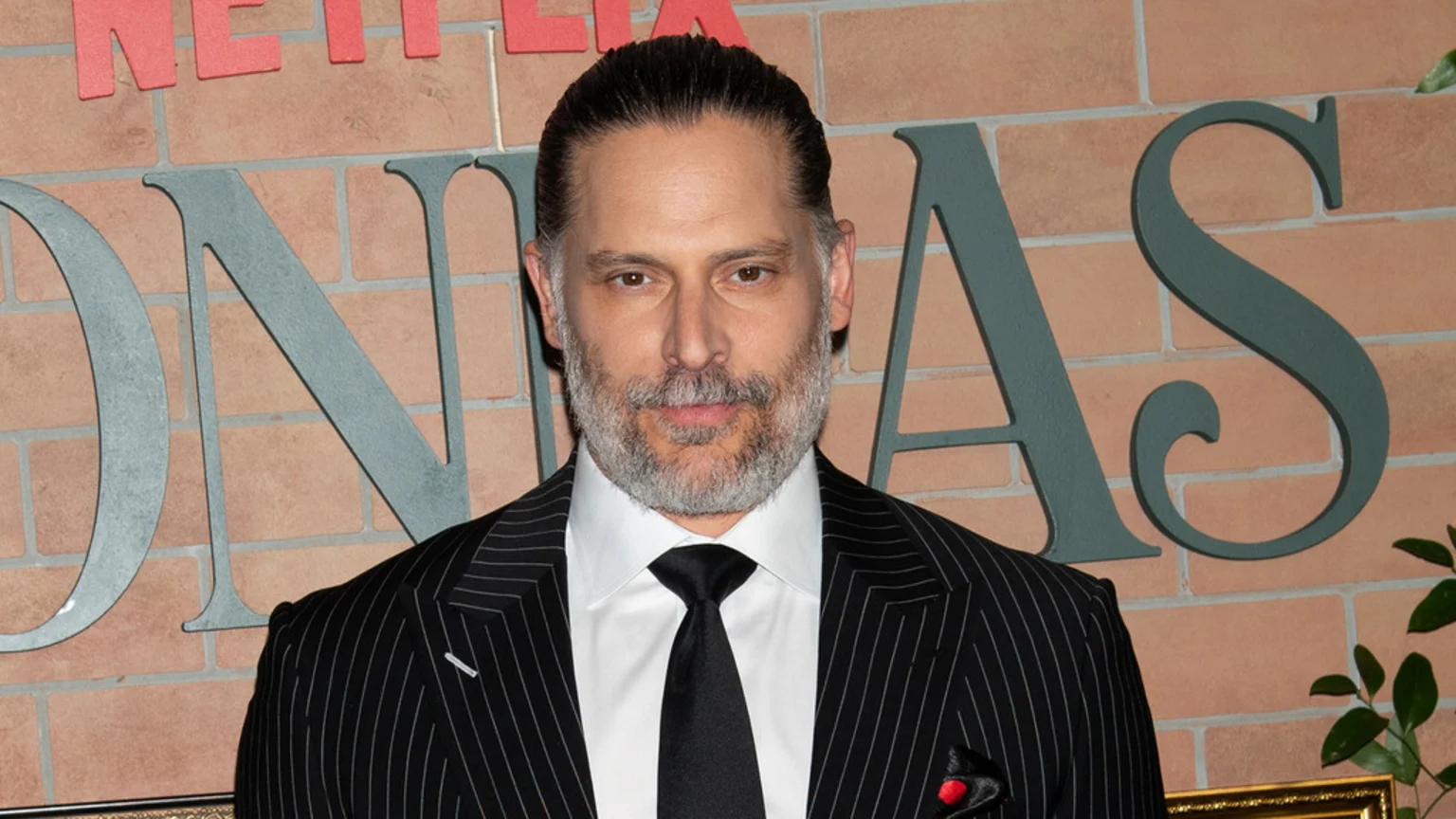 Joe Manganiello with a silver-grey beard and slicked-back dark hair, wearing a black pinstripe suit, white shirt, and black tie, stands in front of a brick wall with large letters "ONAS" and "NETFLI" partially visible.