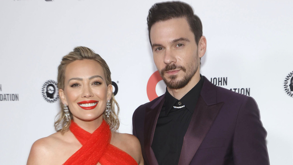 Hilary Duff in a red dress and Matthew Koma in a purple suit stand side-by-side, smiling at the camera against a white background with foundation logos.
