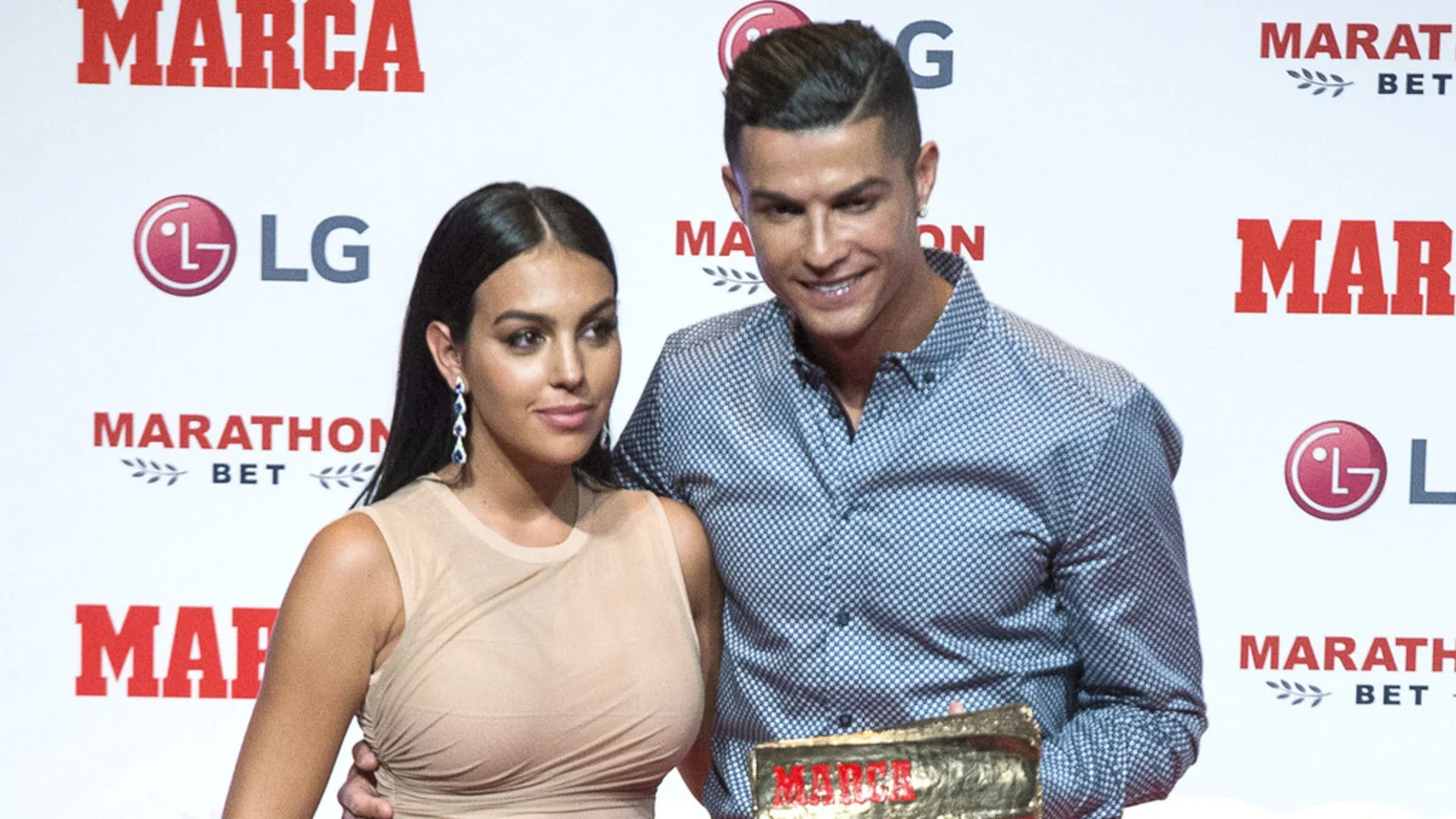 Cristiano Ronaldo and Georgina Rodríguez pose together, with Ronaldo holding a gold award, in front of a white backdrop with "MARCA", "LG", and "MARATHON BET" logos.